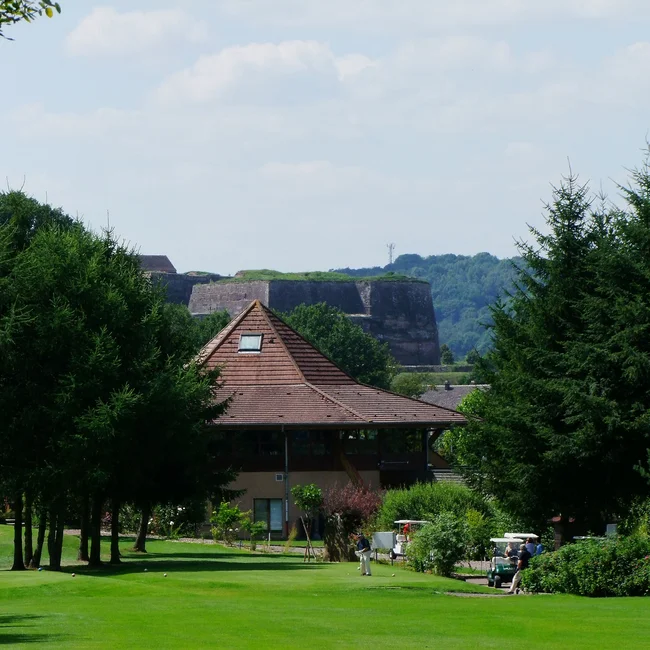 Golfplatz mit Spielern, Bäumen und einem Gebäude mit spitzem Dach im Hintergrund