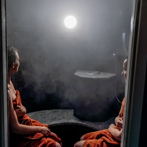 Zwei Frauen in Handtüchern sitzen in einem nebligen Dampfbad