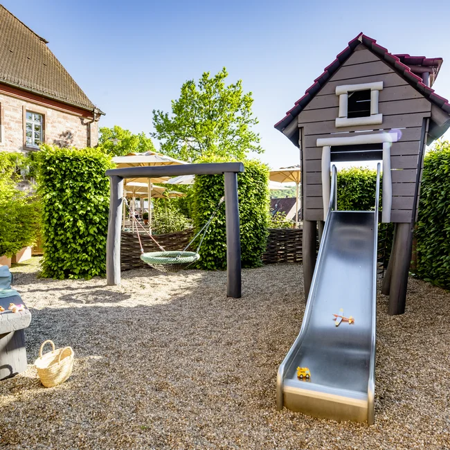 Spielplatz mit Rutsche, Schaukel und Sitzbank neben Haus und grünen Hecken