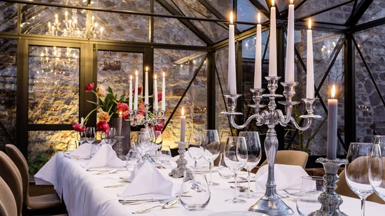 Fein gedeckter Tisch mit Kerzen in elegantem Glasgewächshaus vor Steinwand