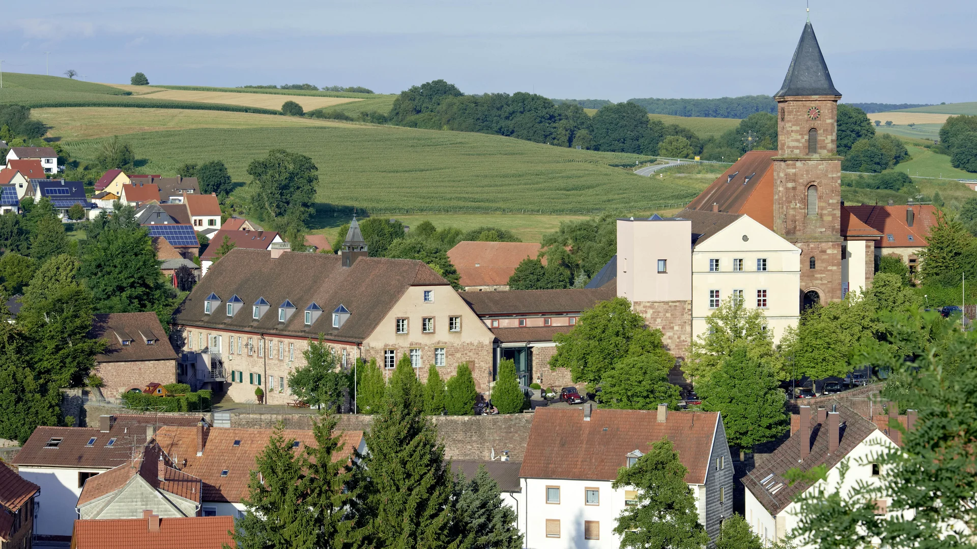 Dorf mit Kirche und Häusern umgeben von grünen Feldern und Hügeln
