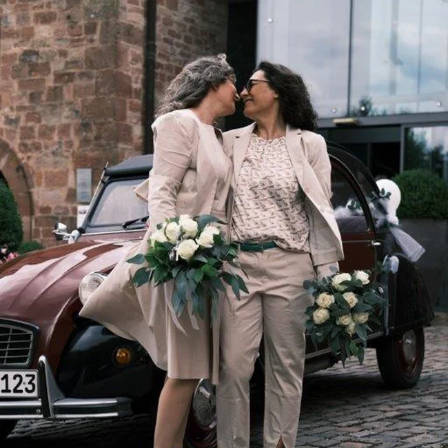 Zwei Frauen im Hochzeitsoutfit stehen vor einem alten Auto, halten Brautsträuße