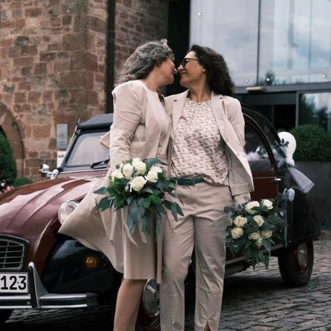 Zwei Frauen im Hochzeitsoutfit stehen vor einem alten Auto, halten Brautsträuße
