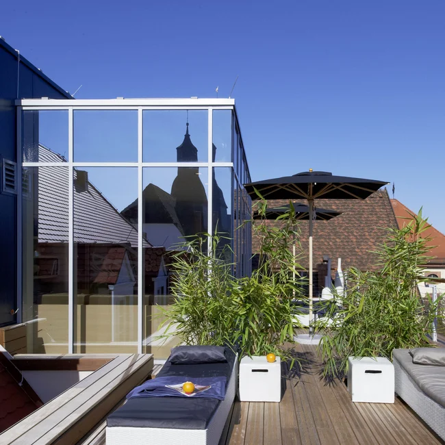 Dachterrasse mit Liegestühlen, Pflanzen und Spiegelung eines Kirchturms im Fenster