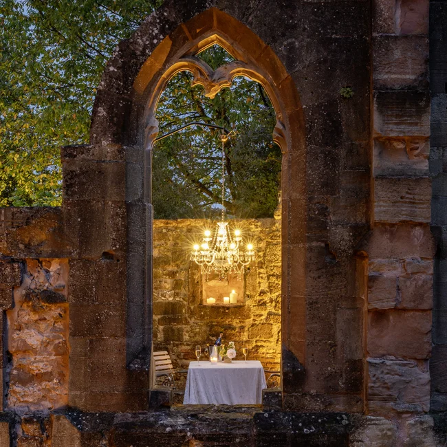 Beleuchteter Esstisch mit Kronleuchter in Ruine mit Steinmauern bei Dämmerung
