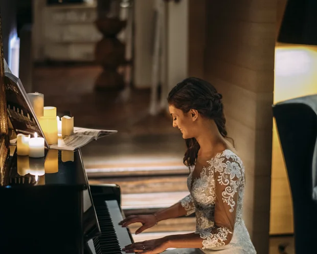 Braut im weißen Kleid spielt bei Kerzenlicht Klavier in gemütlichem Raum