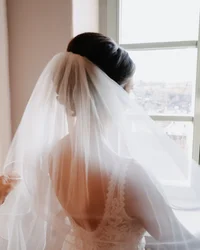 Braut mit Schleier blickt aus Fenster, Hände richten Schleier aus