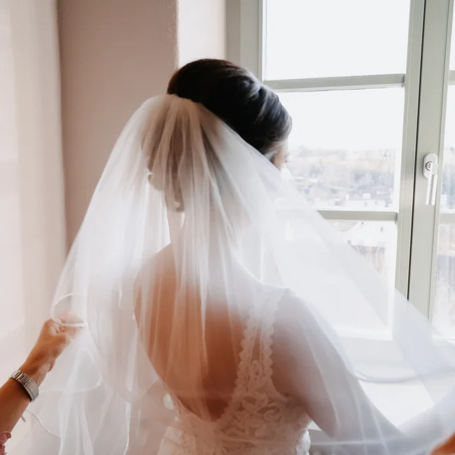 Braut mit Schleier blickt aus Fenster, Hände richten Schleier aus