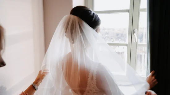 Braut mit Schleier blickt aus Fenster, Hände richten Schleier aus