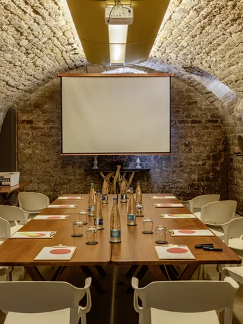 Konferenzraum mit Steintunneldecke, Holztisch, weißen Stühlen und Projektionsleinwand