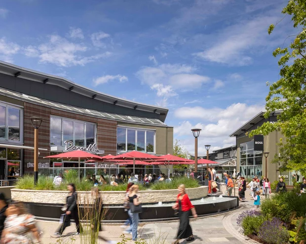 Belebter Einkaufsbereich mit Café und roten Sonnenschirmen unter blauem Himmel