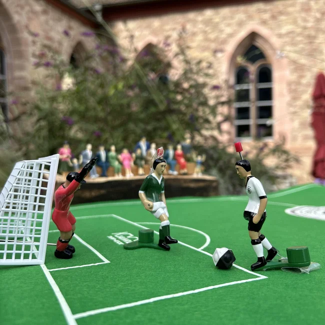 Tischfußballspieler auf grünem Spielfeld vor einem Tor mit Zuschauern im Hintergrund