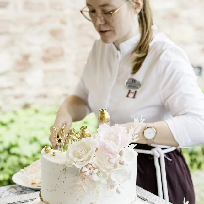 Bäckerin dekoriert elegante weiße Hochzeitstorte mit Blumen und goldenen Verzierungen