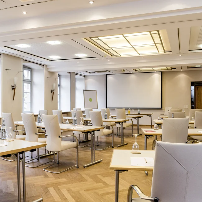Heller Seminarraum mit Tischen, Stühlen, Wasserflaschen und Projektorwand
