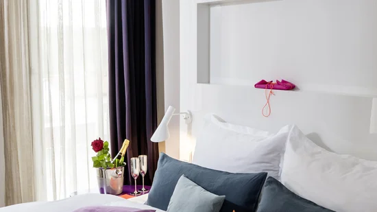 Helles Hotelzimmer mit Bett, lila Decke, bunten Kissen und Champagner mit Rose