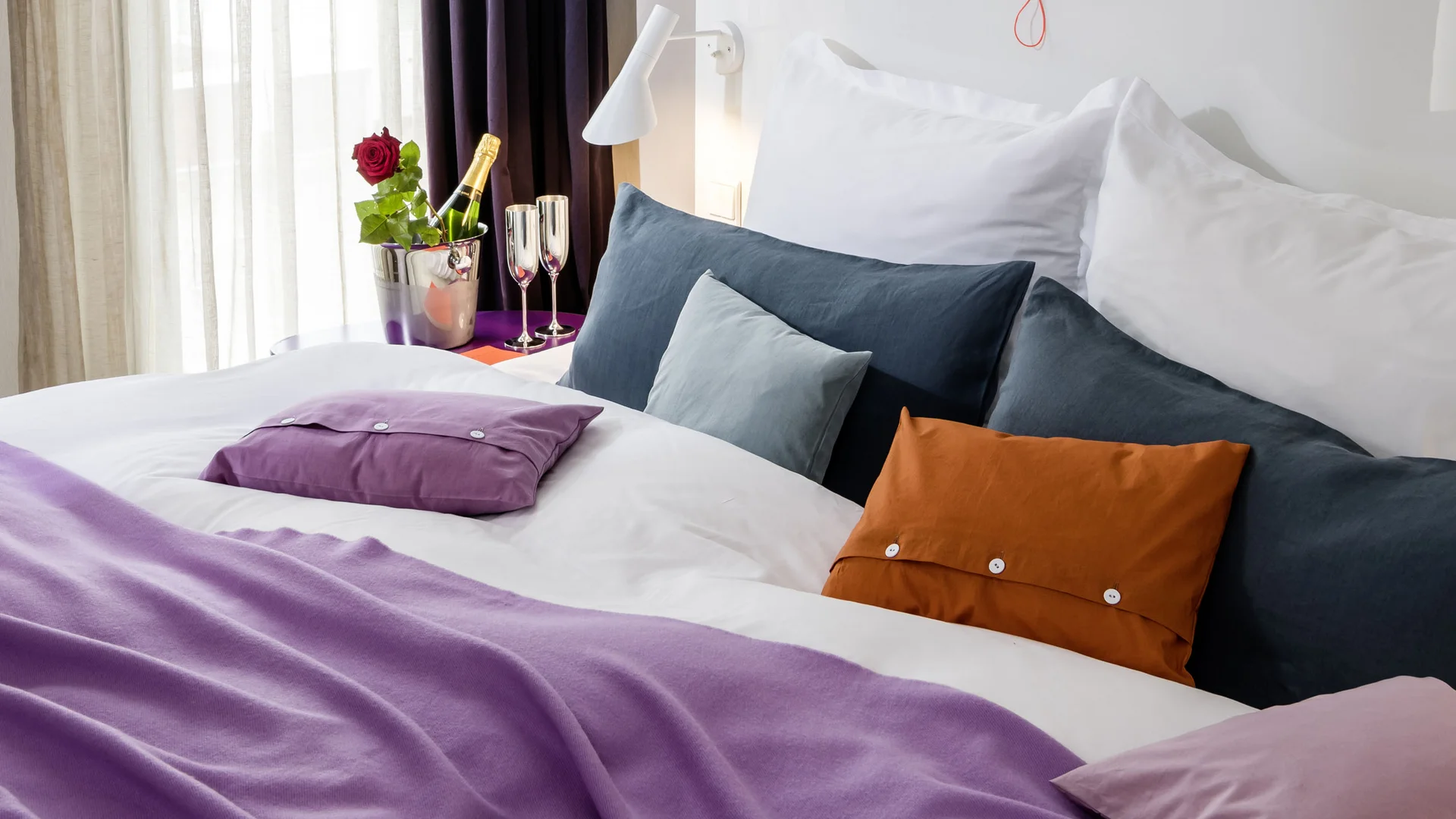 Helles Hotelzimmer mit Bett, lila Decke, bunten Kissen und Champagner mit Rose