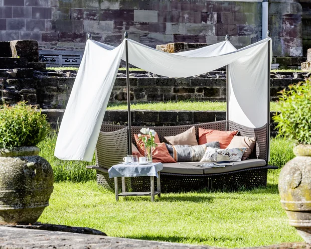 Gemütliche Sitzbank mit Kissen und Sonnenschutz im Garten vor Steinmauer