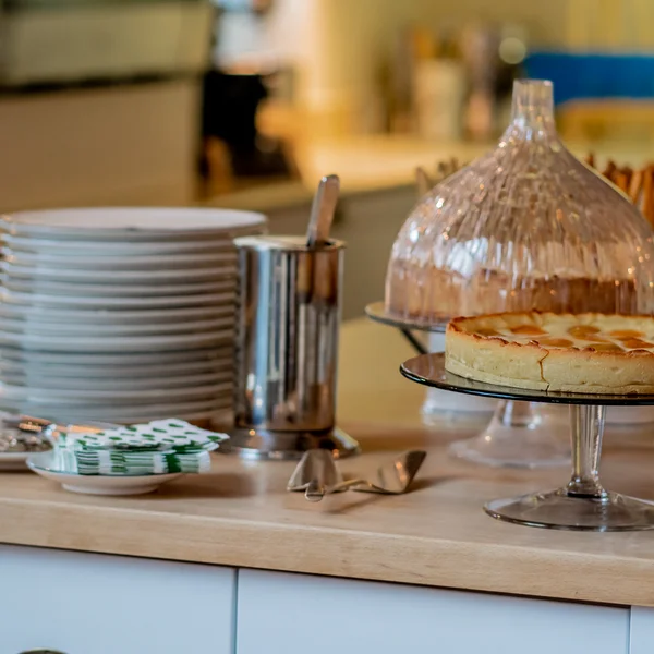 Kuchen auf Glasständer mit Tellern und Besteck auf Holztisch in Küche