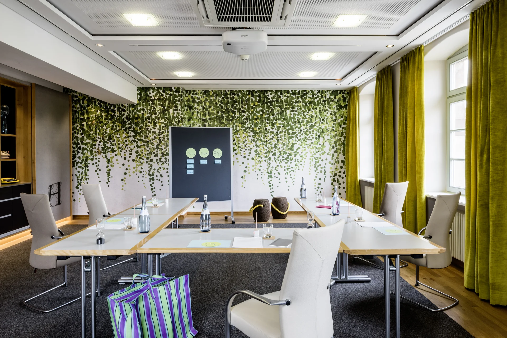 Heller Besprechungsraum mit U-förmigen Tischen, weißen Stühlen und Blätterwanddeko