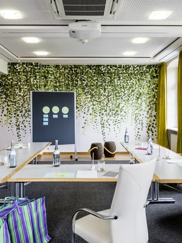 Heller Besprechungsraum mit U-förmigen Tischen, weißen Stühlen und Blätterwanddeko