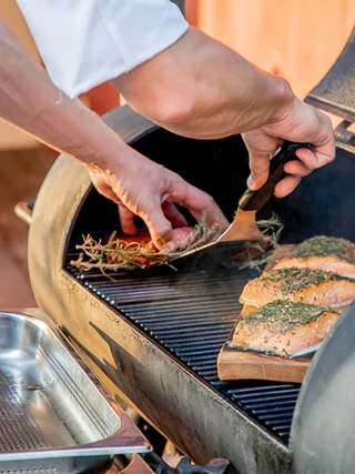 Person legt Lachsfilets mit Kräutern auf einen Grill