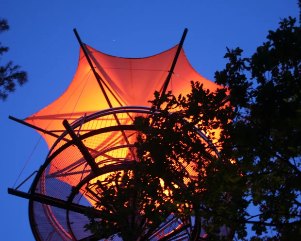 Beleuchteter Aussichtsturm mit oranger Segelstruktur bei Abenddämmerung
