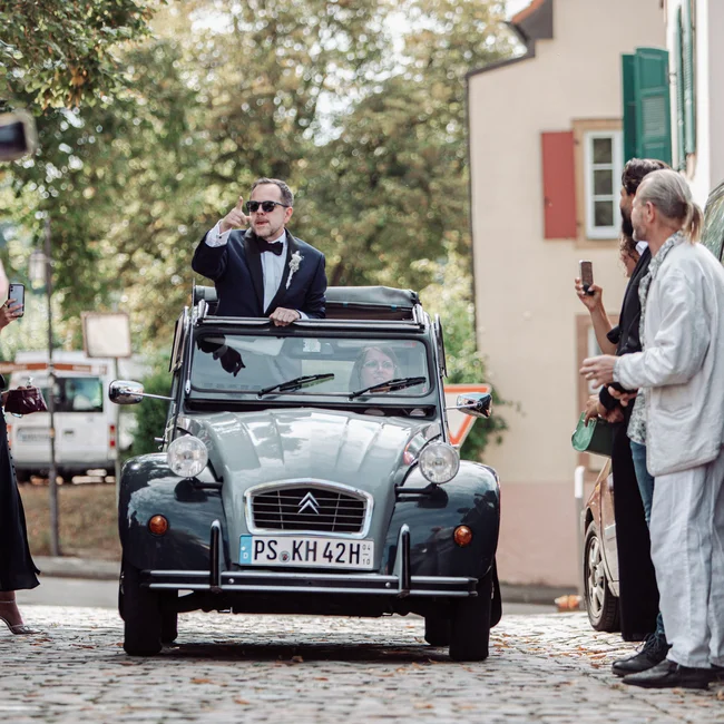 Mann in Anzug winkt aus einem Oldtimer, Leute machen Fotos auf gepflasterter Straße
