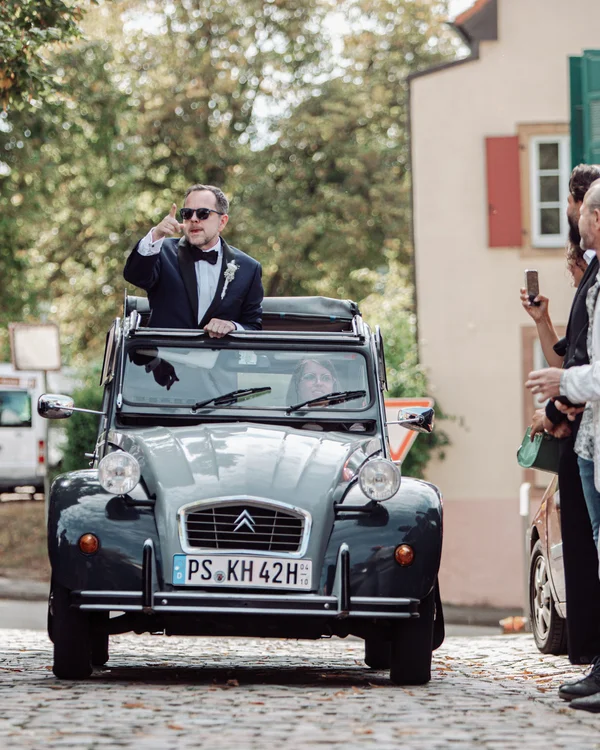 Mann in Anzug winkt aus einem Oldtimer, Leute machen Fotos auf gepflasterter Straße