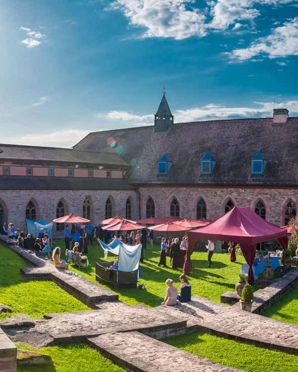 Menschen sammeln sich bei Sonnenschein in einem historischen Innenhof mit Zelten und Sonnenschirmen