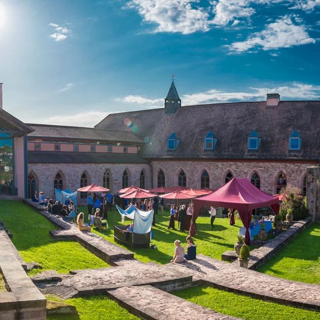 Menschen sammeln sich bei Sonnenschein in einem historischen Innenhof mit Zelten und Sonnenschirmen