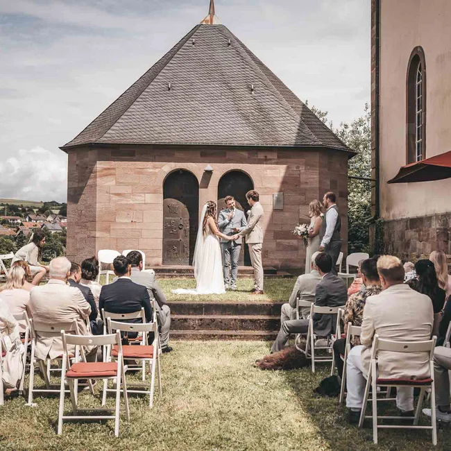 Freilufthochzeit mit Braut, Bräutigam und Gästen vor einem kleinen Steinhaus