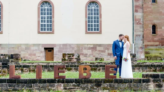 Brautpaar küsst sich neben großen Buchstaben LIEBE auf einer Wiese