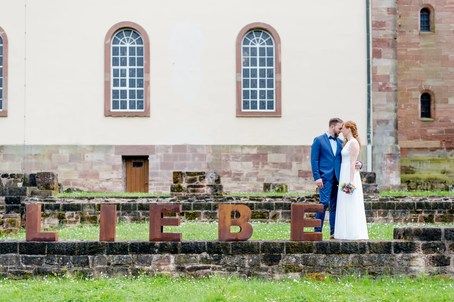Brautpaar küsst sich neben großen Buchstaben LIEBE auf einer Wiese