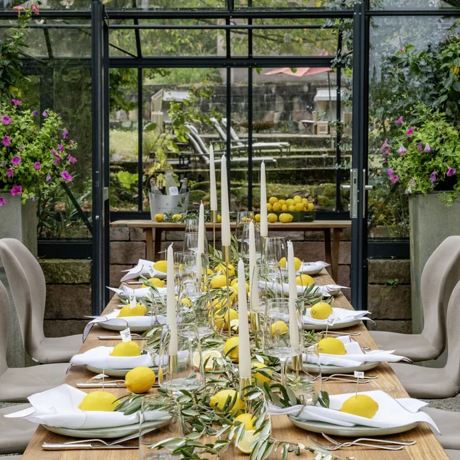Langer Esstisch mit Zitronen, Kerzen und Grün in einem Glashaus