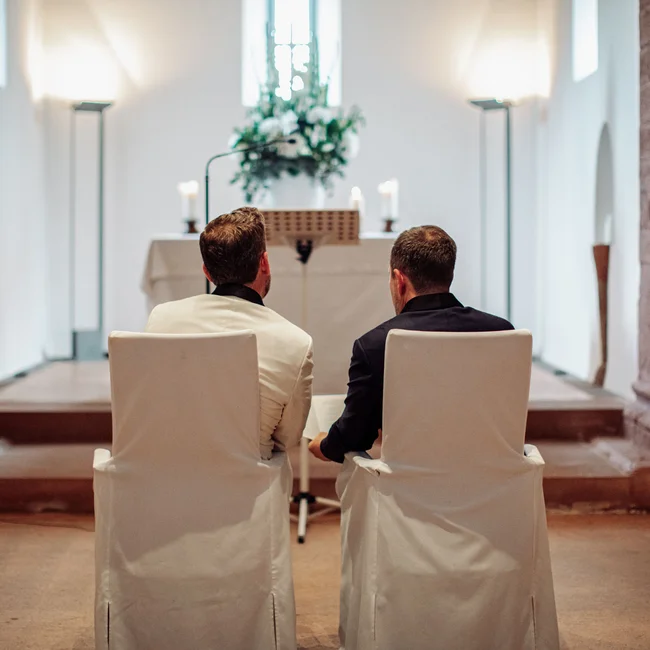 Zwei Männer sitzen in einem festlich geschmückten Raum auf weißen Stühlen