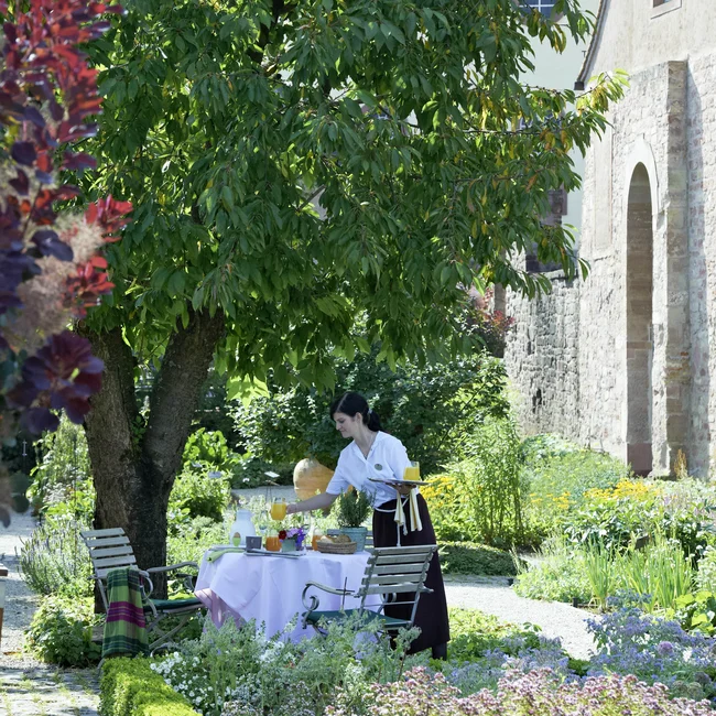 Kellnerin deckt Tisch im Garten neben altem Gebäude an sonnigem Tag
