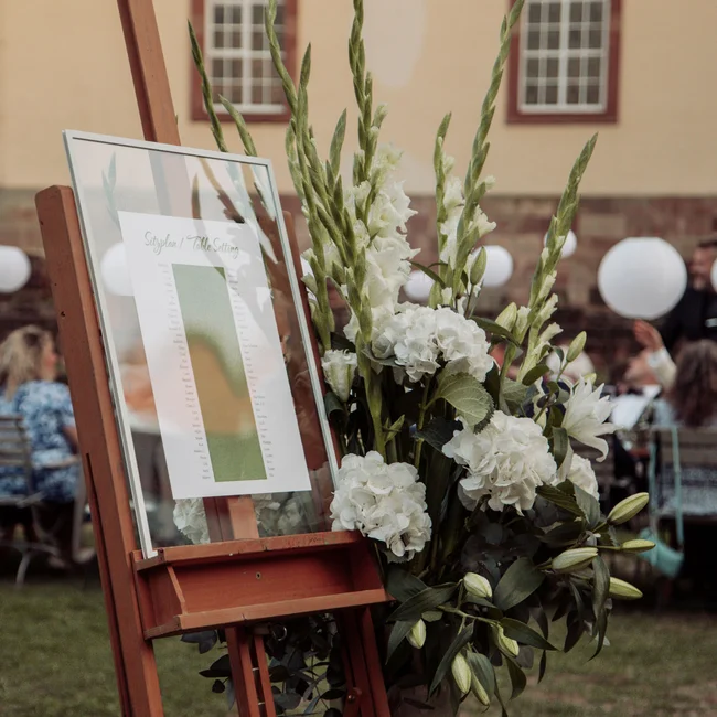Blumenarrangement und Sitzplan auf Staffelei bei Abendveranstaltung im Freien