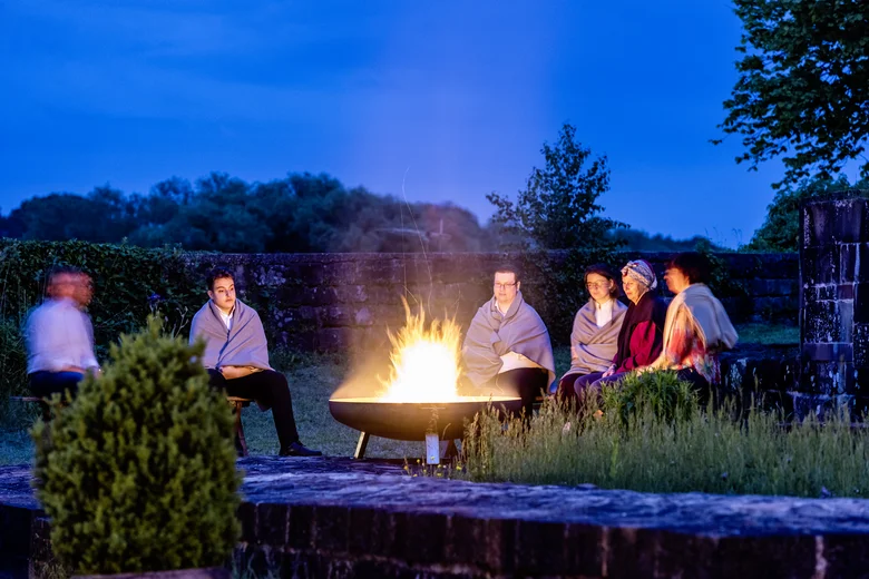 Menschen sitzen abends um ein offenes Feuer im Freien, eingehüllt in Decken