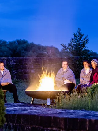 Menschen sitzen abends um ein offenes Feuer im Freien, eingehüllt in Decken