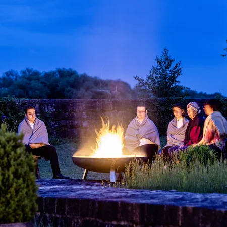 Menschen sitzen abends um ein offenes Feuer im Freien, eingehüllt in Decken