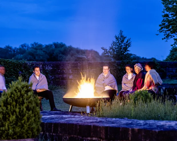 Menschen sitzen abends um ein offenes Feuer im Freien, eingehüllt in Decken