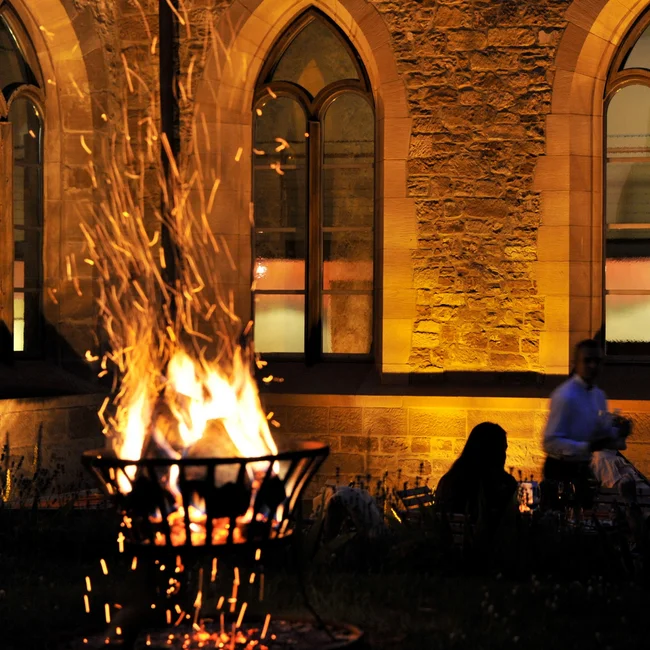 Menschen sitzen bei Feuerkorb vor beleuchtetem steinernen Gebäude bei Nacht