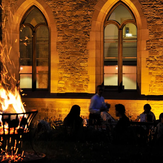 Menschen sitzen bei Feuerkorb vor beleuchtetem steinernen Gebäude bei Nacht
