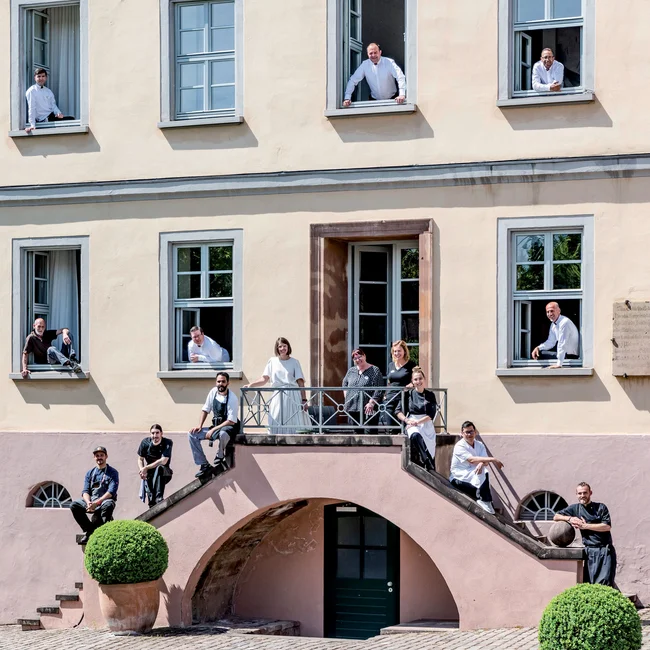 Gruppe von Menschen auf Außentreppe und an Fenstern eines hellen Gebäudes
