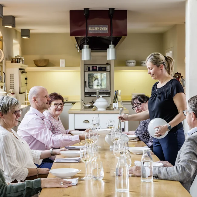 Gruppe älterer Personen am Esstisch in heller Küche, Frau serviert Essen