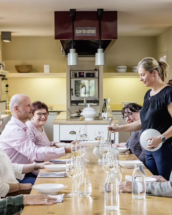 Gruppe älterer Personen am Esstisch in heller Küche, Frau serviert Essen