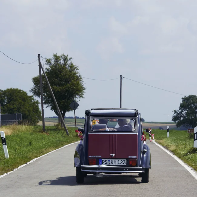 Rotes Retro-Auto fährt auf Landstraße mit grünen Feldern und Bäumen