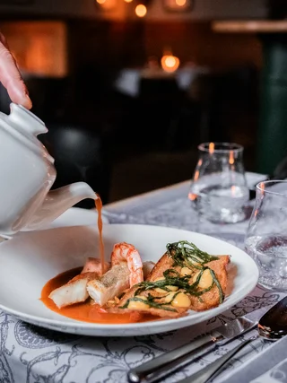 Hand gießt Sauce aus Kanne über Gourmetgericht in weißem Teller im Restaurant