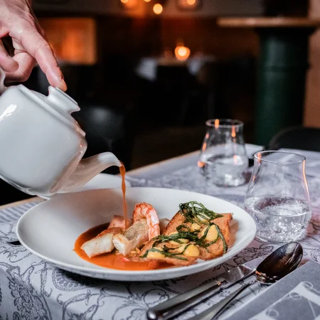 Hand gießt Sauce aus Kanne über Gourmetgericht in weißem Teller im Restaurant