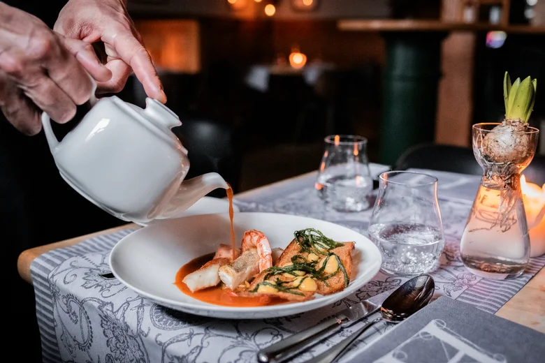 Hand gießt Sauce aus Kanne über Gourmetgericht in weißem Teller im Restaurant